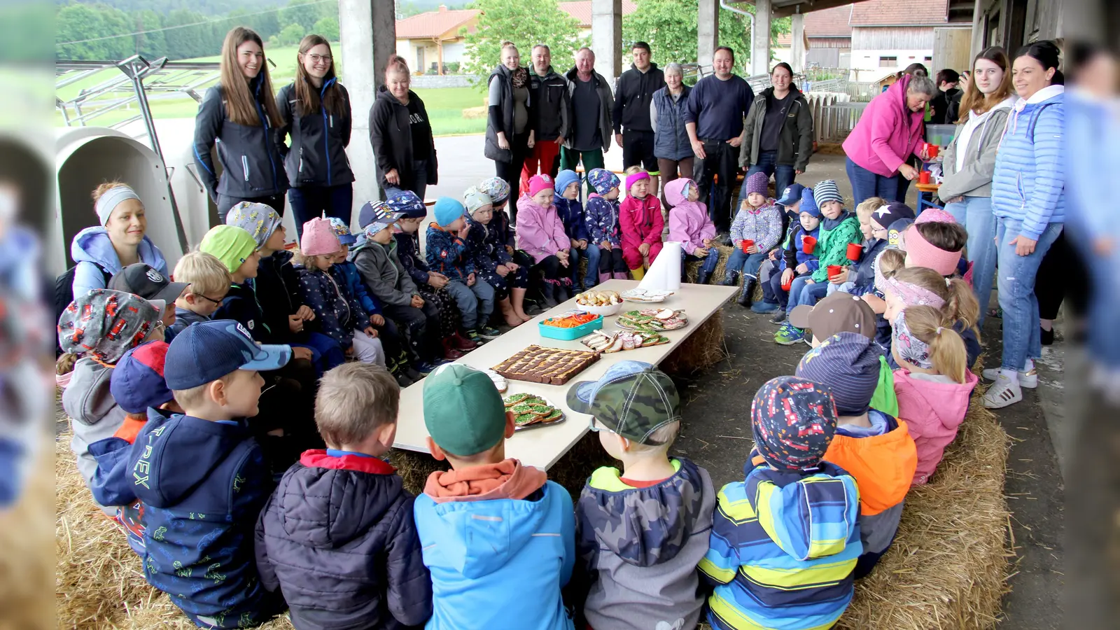 Nach der Besichtigungsrunde durften die Kinder am gedeckten Tisch Platz nehmen. Leckeres vom Bauernhof spendierte die Familie Salomon. Im Bild hinten stehend von rechts die Bauernhofbewirtschafter Maria und Martin Salomon mit ihren Familienangehörigen und dem Auszubildenden; ganz links stehend die bayerische Milchkönigin Elisabeth Heimerl. (Bild: Josef Böhm)
